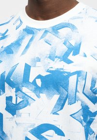 Εγγύς πλάνο ενός λευκού πουκαμίσου με αφηρημένο μπλε σχέδιο ψεκασμού που αποτελείται από επικαλυπτόμενα γράμματα στο στήθος και στους ώμους.