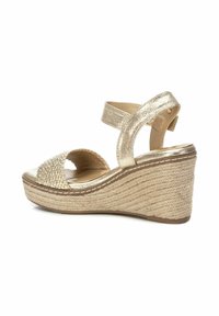 Sandalia cuña dorada con textura trenzada y punta abierta. Presenta correa en el tobillo, tacón de estilo yute trenzado y superficie metálica suave.