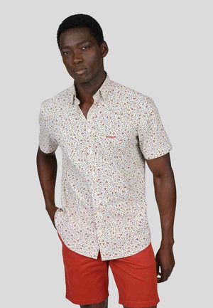 Chemise à manches courtes avec un fond blanc, ornée d'un motif floral multicolore et d'un logo rouge sur la poitrine. Associée à un short rouge.