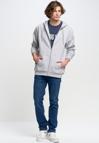 Noor mees seisab käed taskutes, seljas hall lukuga kapuutsiga dressipluus, tumesinine särk, sinised teksad ja valged tossud, ühevärvilise taustaga.