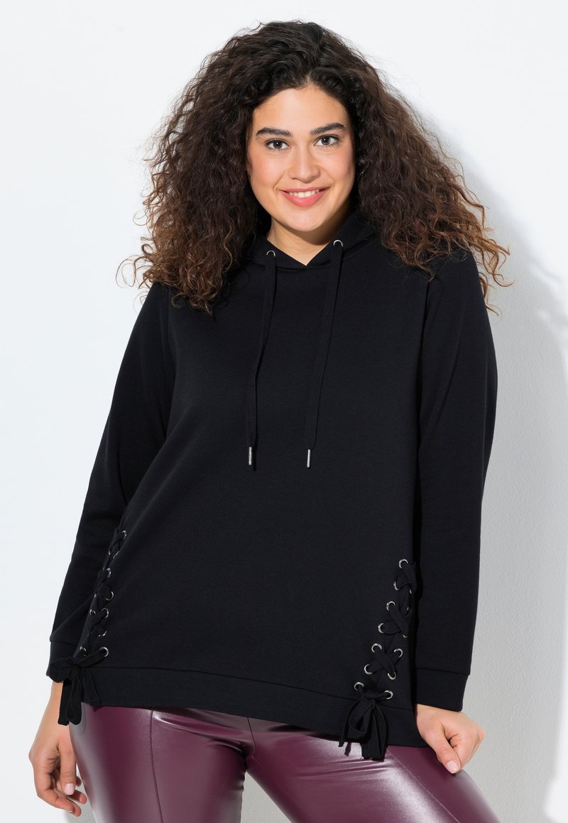 Donna sorridente con capelli ricci che indossa un felpa nera con dettagli in pizzo laterale e pantaloni viola lucidi, in piedi davanti a uno sfondo bianco.