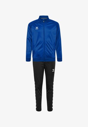 Blaue Sportjacke aus glattem Stoff mit Reißverschluss vorne, kombiniert mit schwarzen Hosen, die Streifen und Logo-Akzente an den Seiten aufweisen.