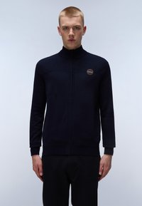 Marinblå zip-up tröja med hög krage, texturerad överdel, ribbad mudd och nederkant. Har en rund logotyp på vänster bröst. Slät stickad tyg.