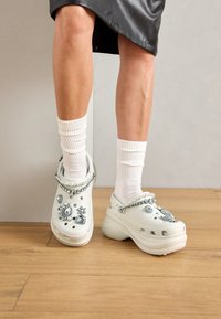 Ben som bär vita strumpor och vita plattformsträskor dekorerade med stora rhinestones och pärlliknande beads på ett trägolv.