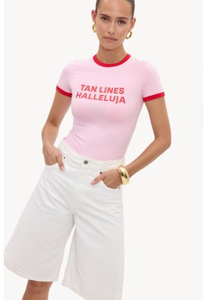 Femme portant un t-shirt rose ajusté avec des bordures rouges et le texte "TAN LINES HALLELUJA,", un short blanc taille haute à jambes larges, ainsi qu'un bracelet et des boucles d'oreilles en or.