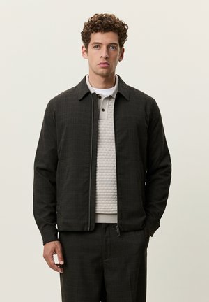 Jeune homme aux cheveux bouclés portant une veste zippée sombre, un pull beige texturé et un pantalon sombre assorti, debout devant un fond uni.