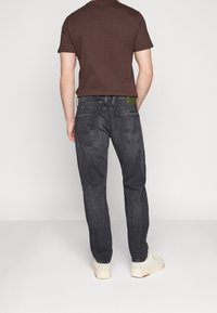 Jeans gris foncé avec une coupe décontractée et une couture subtile sur les poches arrières, associés à un T-shirt marron et des baskets blanches. Détail de l'étiquette verte visible.
