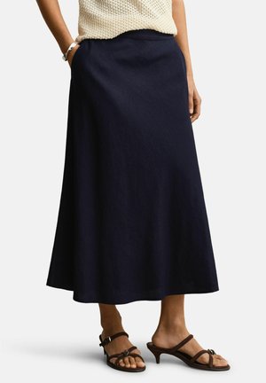 Frau trägt einen dunkelblauen Midirock, ein cremefarbenes ärmelloses Stricktop, ein silbernes Armband und braune Sandalen mit Absatz, steht mit den Händen in den Taschen.