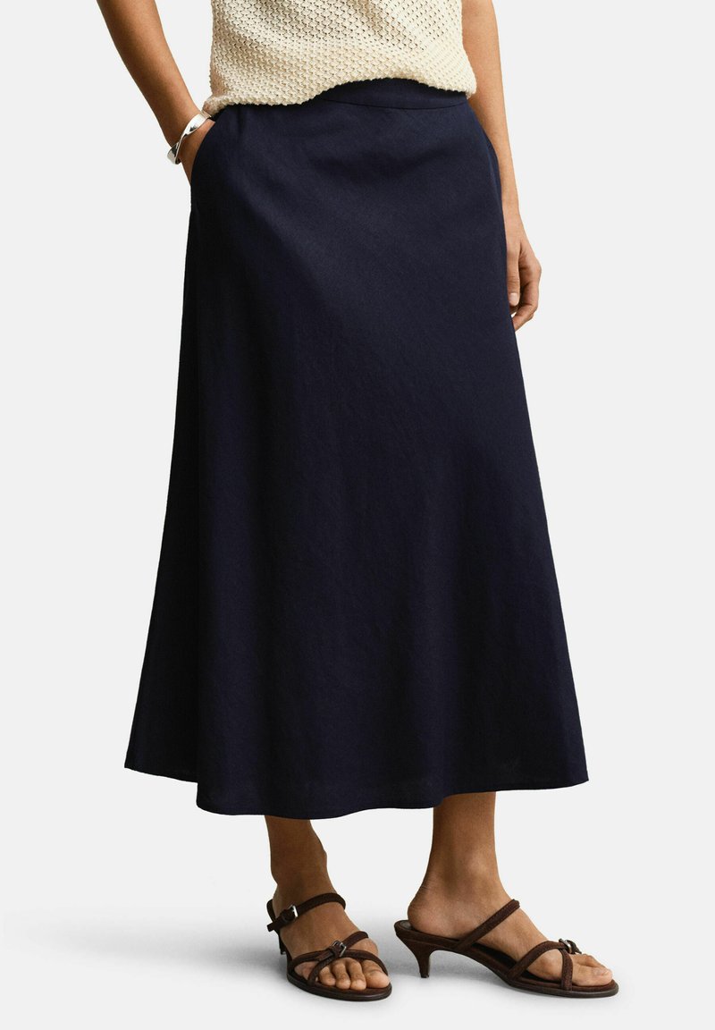 Frau trägt einen dunkelblauen Midirock, ein cremefarbenes ärmelloses Stricktop, ein silbernes Armband und braune Sandalen mit Absatz, steht mit den Händen in den Taschen.