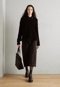 Pull en tricot noir avec col, associé à une longue jupe noire. Le modèle tient un sac fourre-tout noir et porte des bottines noires chunky.