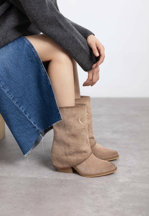 Botines de ante de color beige con tacón bajo, que presentan patrones decorativos de costura; combinados con una falda de mezclilla. Diseño texturizado y casual.