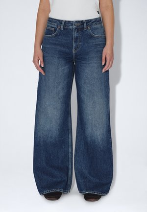 Persona che indossa jeans denim blu scuro a gamba larga e un top bianco, in piedi davanti a uno sfondo chiaro e uniforme.
