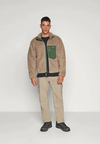 Fleecová bunda v béžové barvě s zelenou náprsní kapsou, zapínání na zip a stojáčkovým límcem, doplněná o světlé béžové kalhoty a černou obuv.