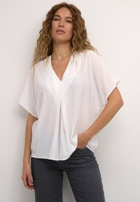 Blusa bianca, ampia e con scollo a V, con maniche corte, realizzata in materiale leggero, caratterizzata da una piega frontale e un drappeggio sottile.