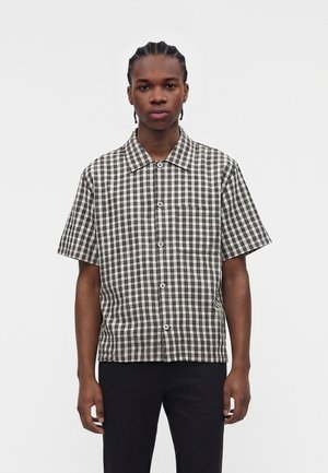 Νεαρό άτομο με κοτσίδες, φοράει κοντομάνικο ασπρόμαυρο πουκαμίσα με καρό μοτίβο και μαύρο παντελόνι, στέκεται μπροστά σε απλό φόντο.