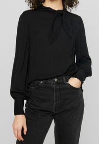 Blusa nera con collo annodato, maniche lunghe a sbuffo e bottoni sulle spalle. Indossata con jeans neri a vita alta. Tessuto morbido e leggero.