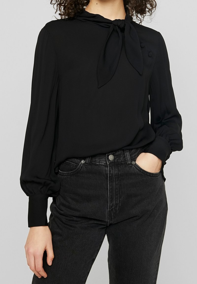 Blusa nera con collo annodato, maniche lunghe a sbuffo e bottoni sulle spalle. Indossata con jeans neri a vita alta. Tessuto morbido e leggero.