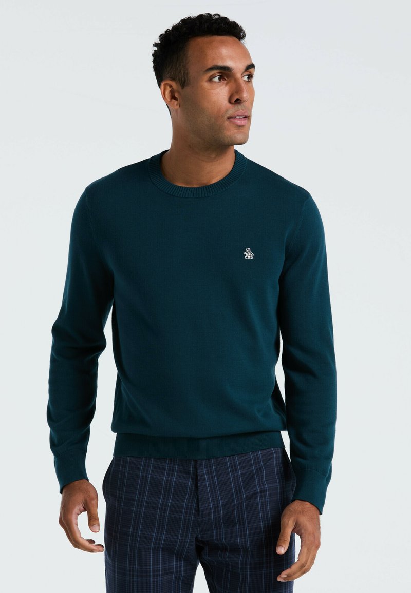 Pull en tricot vert foncé avec un col rond et des poignets côtelés. Présente un petit logo sur la poitrine. Porté avec un pantalon à carreaux bleu marine.