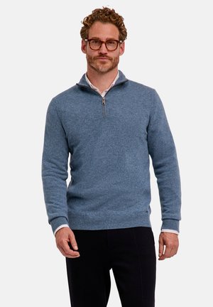 Hombre con cabello rizado y gafas que lleva un jersey azul claro con cremallera parcial sobre una camisa blanca y pantalones negros, de pie frente a un fondo sencillo.
