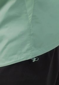 Veste légère vert clair avec une texture lisse, dotée d'un ourlet ajustable avec cordon de serrage et d'accents cousus. Pantalons noirs en dessous.