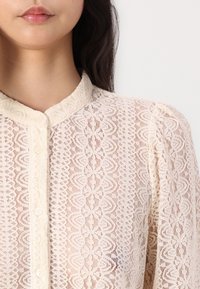 Spetsblus med hög krage, beige färg, med blommönster, knappstängning och lätt veckning vid axlarna.