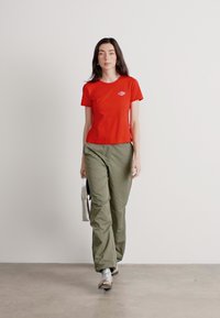 Crvena uska majica s malim logom na prsima, u kombinaciji s širokim maslinastozelenim hlačama i srebrnim sportskim cipelama. Poza u hodu.