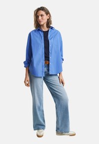 Chemise bleue à boutons avec manches longues portée sur un haut bleu marine, associée à un jean large bleu clair et des baskets blanches.