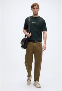 Σκούρο πράσινο κοντομάνικο μπλουζάκι με λευκό λογότυπο, μπεζ παντελόνι χαλαρής εφαρμογής, και αθλητικά παπούτσια ανοιχτού χρώματος. Μαύρη τσάντα χιαστί με λουράκι.