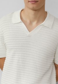 Polo blanc à manches courtes avec un col en V, motif en tricot texturé et col contrasté. Le tissu semble léger et respirant.