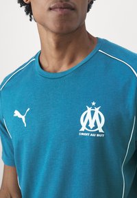 T-shirt in cotone turchese con dettagli bianchi, dotato di un logo Puma sul lato sinistro del petto e di un logo del Marsiglia con testo sottostante. Collo rotondo.