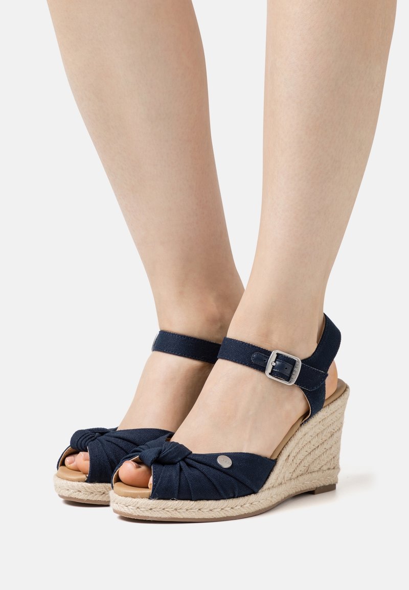 Pieds portant des sandales compensées en tissu bleu marine avec des brides à la cheville et des talons en jute tressé sur un fond blanc uni.