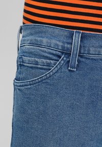 Blå denimjeans med en slät yta, med en framficka, en metallknapp och dragkedjestängning, kompletterad av en randig orange och svart tröja.