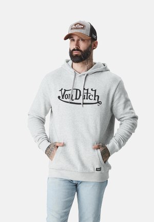 Homme avec barbe et tatouages portant un sweat à capuche gris Von Dutch et une casquette, debout les mains dans les poches du sweat à capuche sur un fond uni.