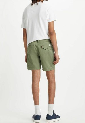 Homme portant un t-shirt blanc, un short vert olive avec une poche arrière à bouton, des chaussettes blanches et des baskets bleu marine, se tenant de dos.