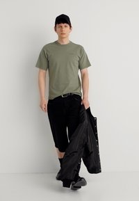 Νεαρός άνδρας με μαύρο καπέλο, ελαι πράσινο μπλουζάκι, μαύρο σορτς και παπούτσια, κρατώντας ένα μαύρο μπουφάν στο δεξί χέρι ενώ περπατά.