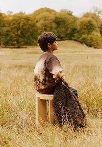 Bambino che indossa un maglione marrone con una grafica di orso, seduto su uno sgabello di legno chiaro e tiene in mano una coperta scura e texturizzata.