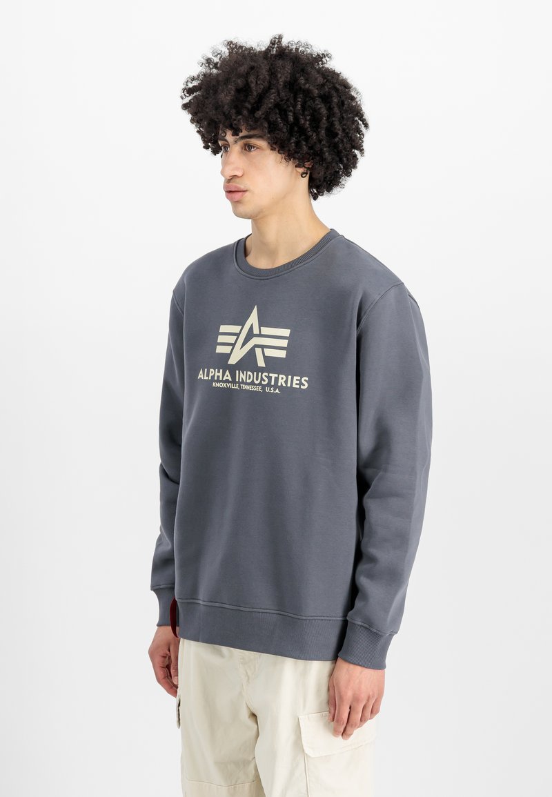 Sudadera gris hecha de tejido suave, con un gran logo blanco en el pecho, cuello redondo y puños y dobladillo de canalé. El modelo lleva pantalones cortos claros.