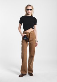 Maglietta nera cropped, pantaloni ampi marroni a vita alta e occhiali da sole scuri. Una mano tiene una sciarpa multicolore. Scarpe marroni completano l'outfit.