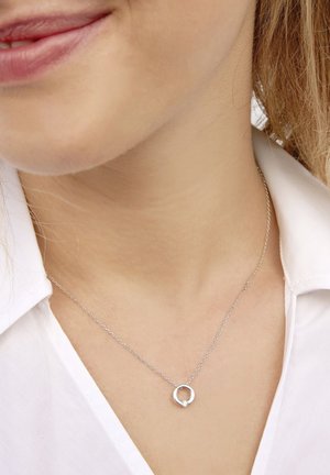 Collana in argento con un pendente circolare e un piccoloAccent di pietra preziosa. La catena è delicata e sembra essere realizzata in metallo di alta qualità. Indossata sopra una camicia bianca.