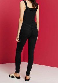 Vaqueros negros ajustados con cintura alta, textura suave y bolsillos traseros. Combinados con sandalias cruzadas negras y crema sobre un fondo rosa.