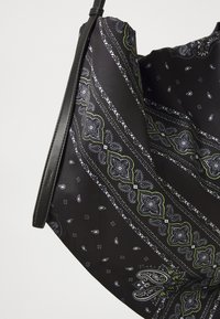 Melna rokassoma ar detalizētu paisley rakstu, kas izceļas ar baltām un zaļām akcentiem, kā arī plānu melnu ādas siksnu.