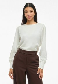 Kvinna med en vit långärmad blus med smockade ärmslut och höga mörkbruna byxor, stående mot en vit bakgrund.