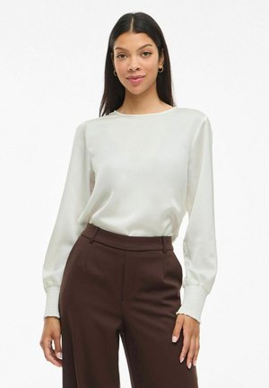 Woman wearing a white long-sleeve blouse with smocked cuffs and high-waisted dark brown pants, standing against a white background.