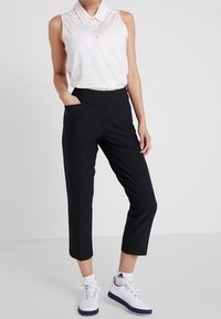 Kvinna i svarta cropped byxor, vit ärmlös blus med krage och ljusblommönster, vita strumpor och vita sportskor som står mot en enfärgad bakgrund.