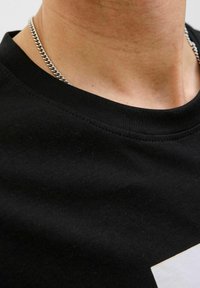 Camiseta de algodón negra con escote redondo, textura suave y un diseño gráfico blanco en la parte frontal. Cadena plateada visible.