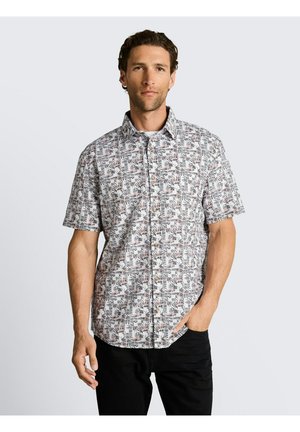 Mężczyzna w krótkim rękawem, zapinanej na guziki koszuli z czarno-brązowym wzorem liści, w połączeniu z czarnymi spodniami, stojący na jasnym tle.