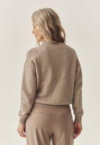 Beige pulóver strukturált chevron mintával, bordás gallérral és mandzsettával, bézs nadrág felett viselve. Nézet hátulról, kiemelve a dizájnt.