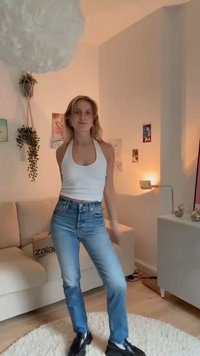 Top branco sem mangas, jeans azuis de cintura alta e sapatos pretos. Interior de uma sala com luz suave, um tapete branco e uma planta.