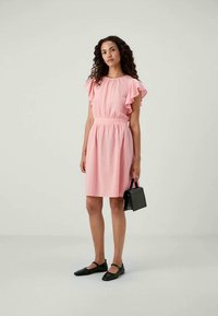 Vestido curto cor-de-rosa com mangas em babados, cintura elástica e um corte descontraído. Usado com sapatos pretos de sola plana e uma pequena bolsa preta.