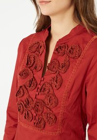 Blusa de algodón roja con cuello alto, aplicaciones decorativas de rosa de crochet y detalles de botones. Ribete de encaje horizontal a lo largo de los bordes.